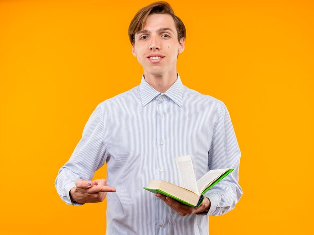 Jonge man in wit overhemd met open boek op zoek glimlachend zelfverzekerd wijzend met wijsvinger op boek staande over oranje muur