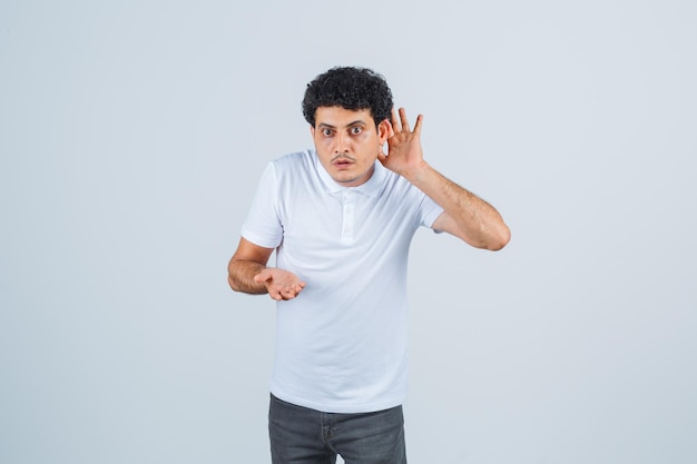 Jonge man in wit t-shirt, broek die hand achter het oor houdt en nieuwsgierig kijkt, vooraanzicht.