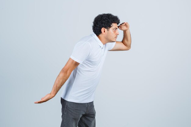 Jonge man in wit t-shirt, broek die ver weg kijkt met de hand boven het hoofd en gefocust, vooraanzicht.