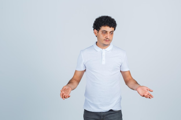 Jonge man in wit t-shirt, broek met hulpeloos gebaar en peinzend, vooraanzicht.