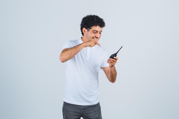 Jonge man in wit t-shirt, broek wijzend op politie walkie talkie telefoon en vrolijk kijkend, vooraanzicht.