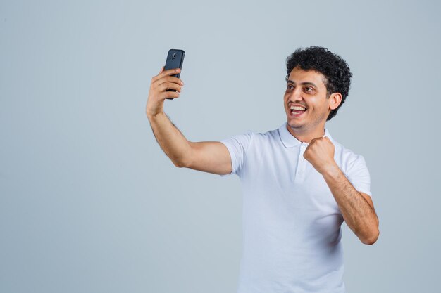 Jonge man in wit t-shirt die naar mobiele telefoon kijkt en er gelukkig uitziet, vooraanzicht.