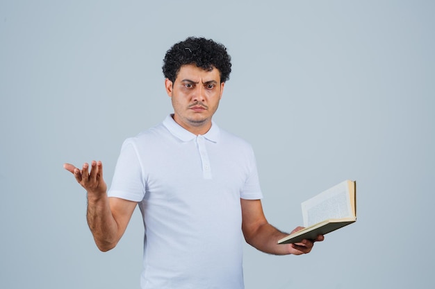 Jonge man in wit t-shirt en spijkerbroek die de hand op een vragende manier uitrekt terwijl hij een boek vasthoudt en verbijsterd kijkt, vooraanzicht.