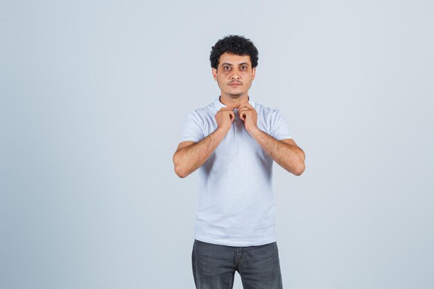 Jonge man in wit t-shirt en spijkerbroek die handen op de kraag zet en er serieus uitziet, vooraanzicht.