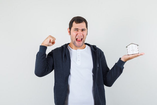 Jonge man in wit t-shirt en zwarte hoodie met rits aan de voorkant met huismodel in de ene hand, spieren tonend en optimistisch op zoek, vooraanzicht.