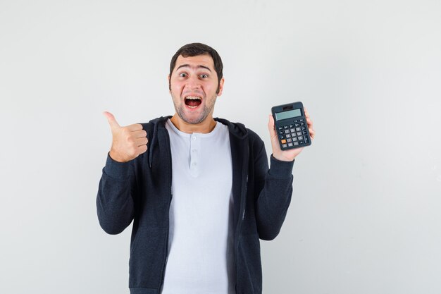 Jonge man in wit t-shirt en zwarte hoodie met rits aan de voorkant met rekenmachine en duim opdagen en op zoek naar optimistisch, vooraanzicht.