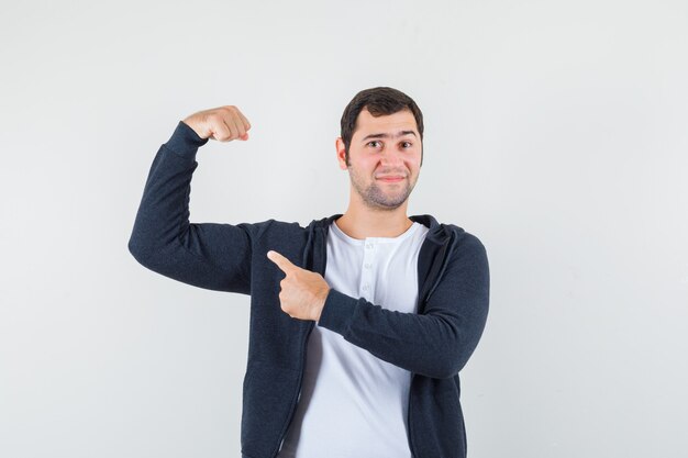 Jonge man in wit t-shirt en zwarte hoodie met ritssluiting die spieren met wijsvinger richt en optimistisch, vooraanzicht kijkt.