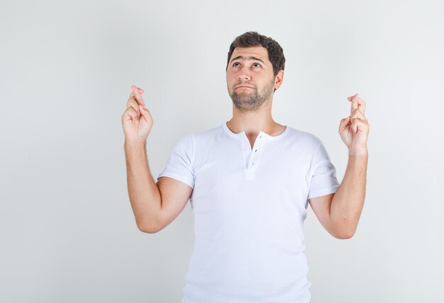 Jonge man in wit t-shirt met gekruiste vingers opzoeken en hoopvol kijken