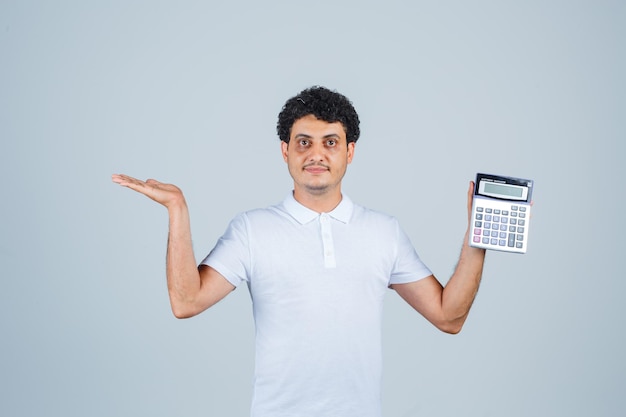 Jonge man in wit t-shirt met rekenmachine terwijl hij de palm opzij spreidt en er zelfverzekerd uitziet, vooraanzicht.