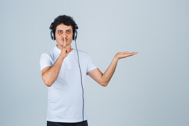 Jonge man in wit t-shirt met stiltegebaar, palm opzij spreidend en verstandig, vooraanzicht.