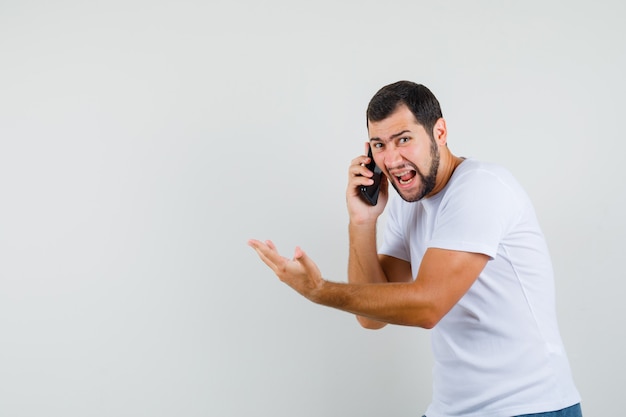 Jonge man in wit t-shirt schreeuwen tijdens het praten aan de telefoon en kijkt boos, vooraanzicht. ruimte voor tekst