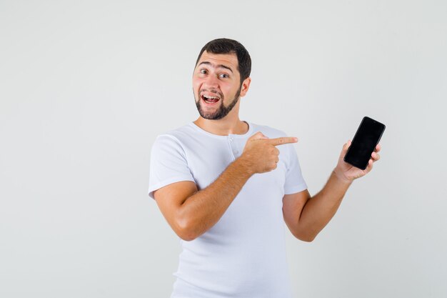 Jonge man in wit t-shirt wijzend op mobiele telefoon en kijkt vrolijk, vooraanzicht.