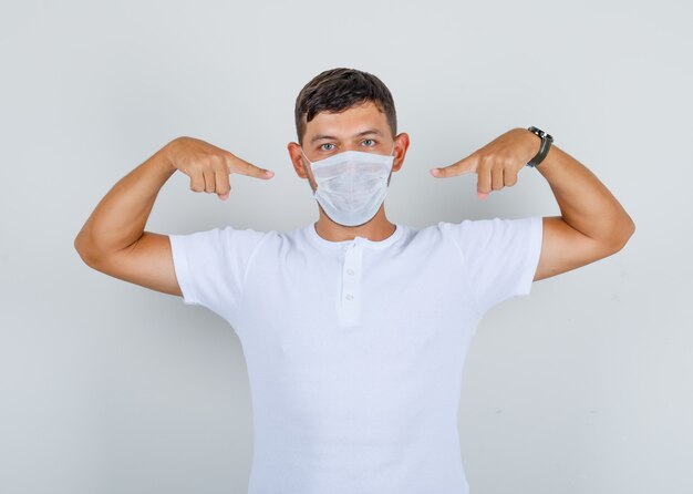 Jonge man in wit t-shirt wijzende vingers op medisch masker, vooraanzicht.