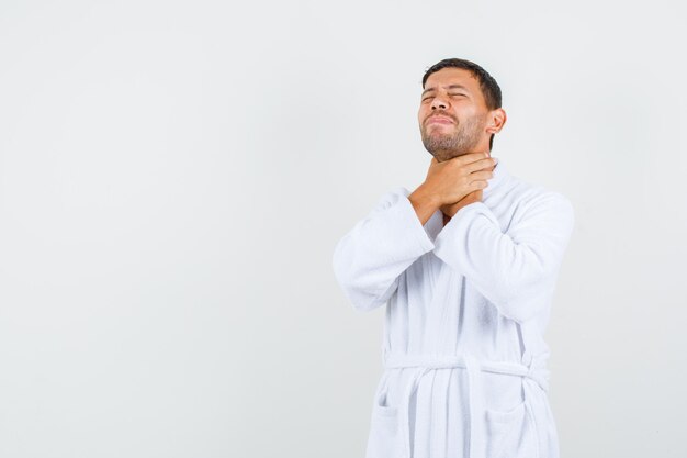 Jonge man in witte badjas hand in hand op zijn keel en op zoek pijnlijk, vooraanzicht.