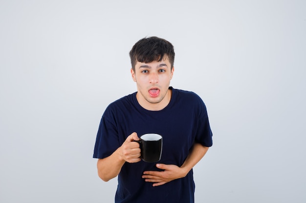 Jonge man in zwart t-shirt misselijk voelen terwijl hij een kopje thee vasthoudt en er onwel uitziet, vooraanzicht.