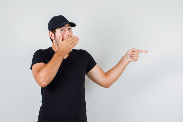 Jonge man in zwart t-shirt, pet naar kant met hand op mond en verbaasd kijken
