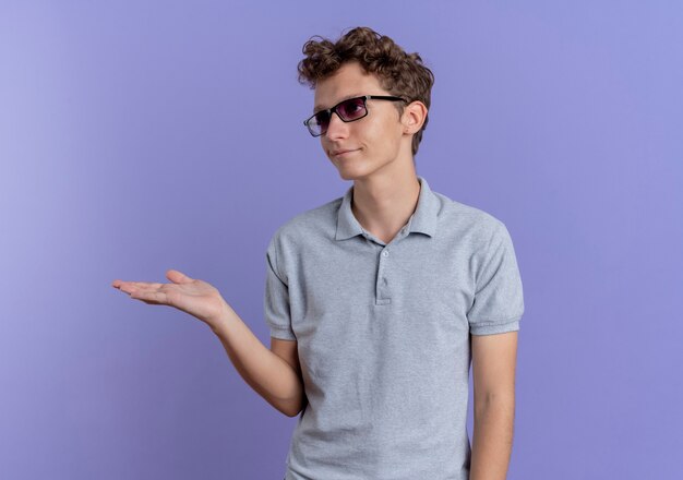 Jonge man in zwarte bril die grijs poloshirt draagt dat iets met handarm voorstelt die met sceptische uitdrukking over blauwe muur kijkt