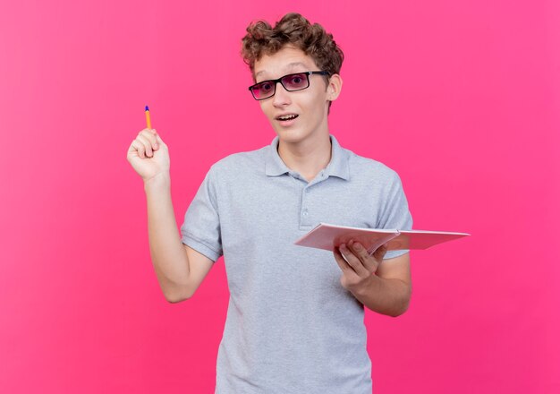 Jonge man in zwarte bril die grijs poloshirt draagt die notitie met pen houdt die blij en opgewonden glimlacht met nieuw idee dat zich over roze muur bevindt