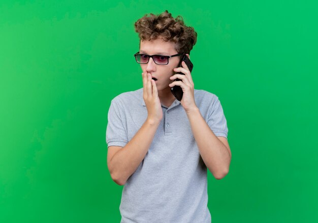Jonge man in zwarte bril dragen grijze poloshirt praten op mobiele telefoon wordt verbaasd en verrast over groen