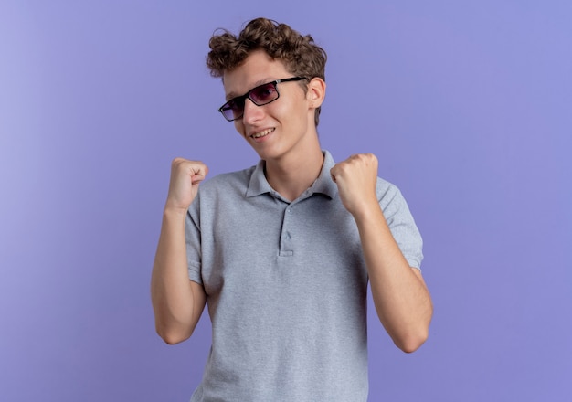 Jonge man in zwarte bril, gekleed in een grijs poloshirt met gebalde vuisten blij en opgewonden staande over blauwe muur