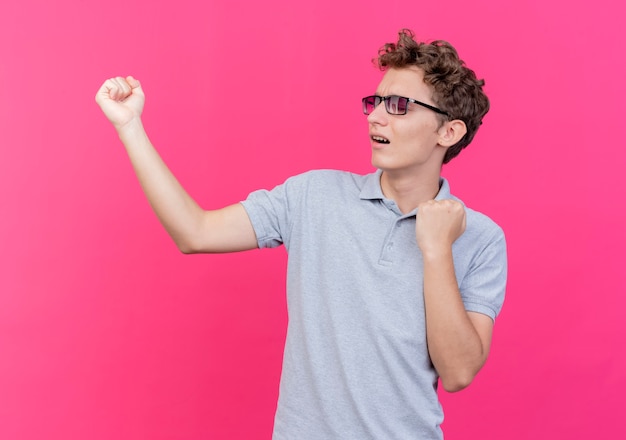 Jonge man in zwarte bril, gekleed in grijs poloshirt balde vuisten blij en opgewonden over roze