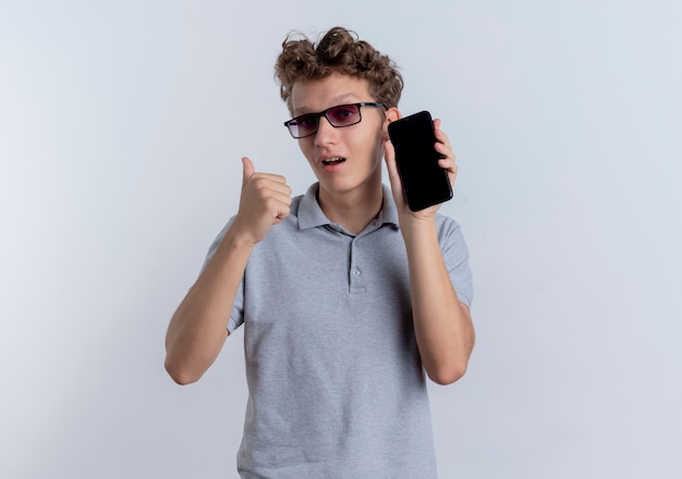 Jonge man in zwarte bril, gekleed in grijs poloshirt met smartphone die terug wijst op zoek verward staande over witte muur