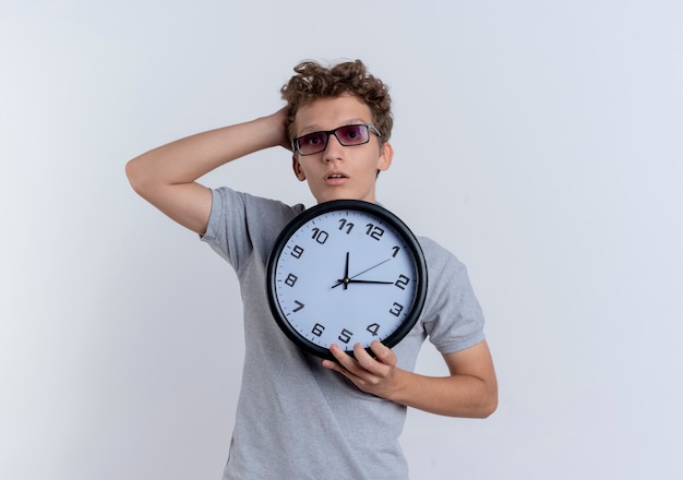 Jonge man in zwarte bril, gekleed in grijs poloshirt met wandklok verward met hand op zijn hoofd staande over witte muur