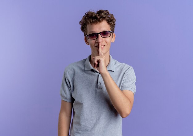 Jonge man in zwarte bril met grijs poloshirt met ernstig gezicht stilte gebaar maken met vinger op lippen staande over blauwe muur