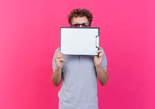 Jonge man in zwarte bril met grijs poloshirt met klembord met blanco pagina's die zijn gezicht erachter verbergen gluren over staande over roze muur