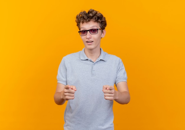 Jonge man in zwarte bril met grijs poloshirt op zoek zelfverzekerd wijzend met wijsvingers blij en positief staande over oranje muur