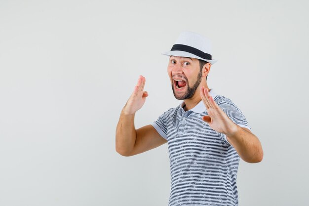 Jonge man karate hakken gebaar in t-shirt, hoed tonen en op zoek zelfverzekerd, vooraanzicht.