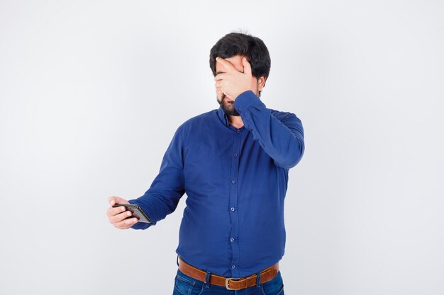 Jonge man kijkt naar de telefoon met de hand op de ogen in een koningsblauw shirt en kijkt bang, vooraanzicht.