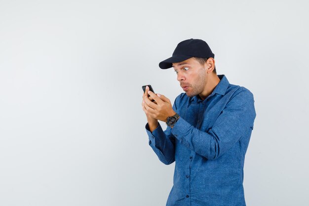 Jonge man kijkt naar mobiele telefoon in blauw shirt, pet en kijkt geschokt. vooraanzicht.