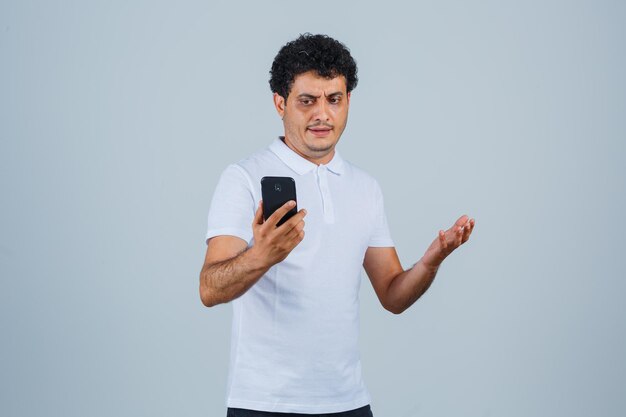 Jonge man kijkt naar mobiele telefoon in wit t-shirt en kijkt teleurgesteld, vooraanzicht.