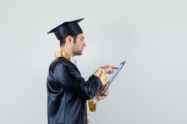 Jonge man kijkt uit over notities op het klembord in afgestudeerde uniform en kijkt bezig.