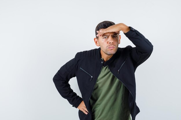 Jonge man kijkt ver weg met de hand boven het hoofd in t-shirt, jas en kijkt peinzend, vooraanzicht.