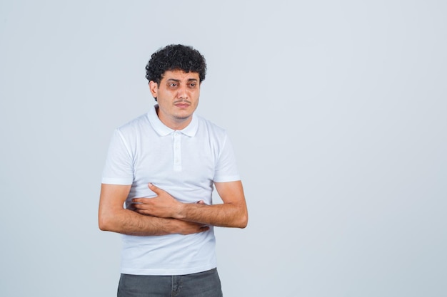 Jonge man knuffelt zichzelf terwijl hij in wit t-shirt, broek denkt en boos kijkt, vooraanzicht.