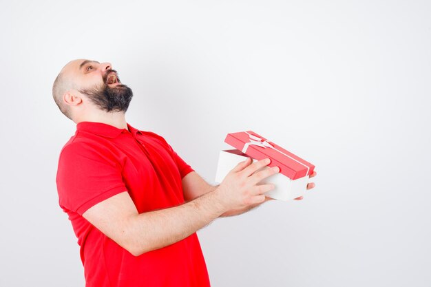 Jonge man lacht terwijl hij een geschenkdoos in een rood t-shirt vasthoudt en er gelukkig uitziet, vooraanzicht.