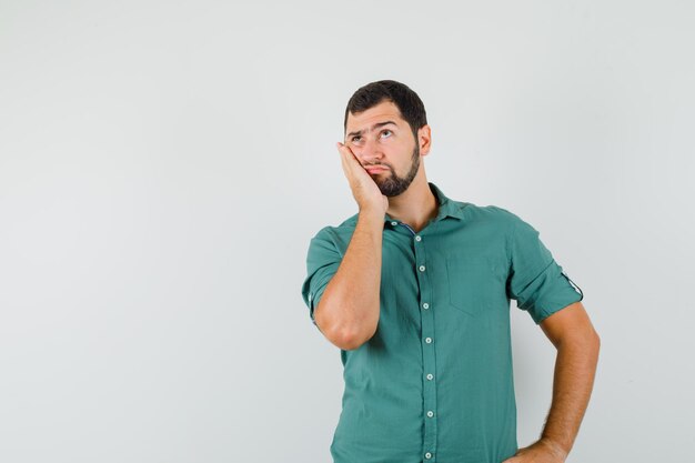 Jonge man leunt op zijn handpalm terwijl hij wegkijkt in een groen shirt en er attent uitziet, vooraanzicht. ruimte voor tekst