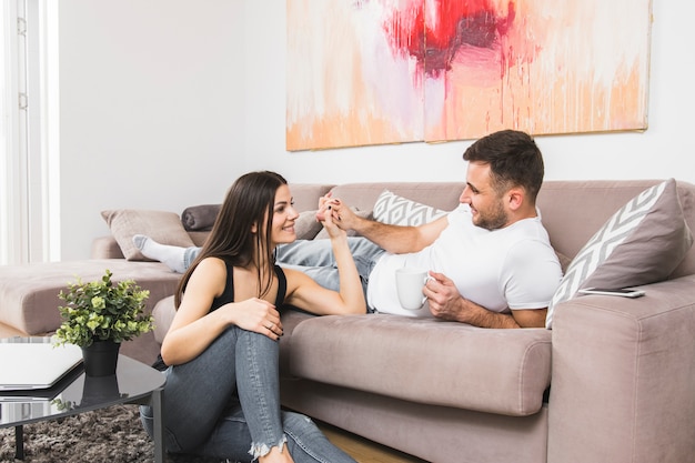 Jonge man liggend op de bank holding hand van zijn vriendin zittend op de grond