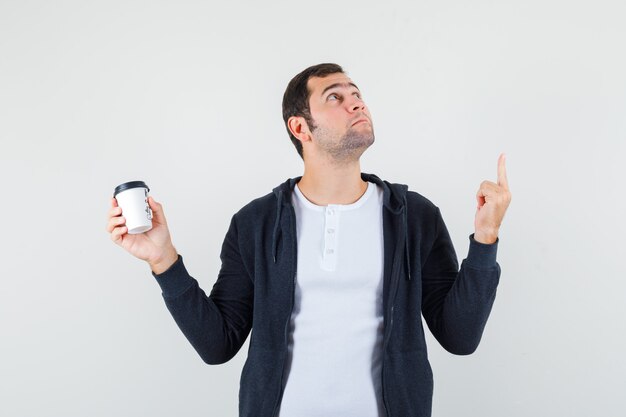 Jonge man met afhaalmaaltijden koffiekopje en omhoog met wijsvinger in wit t-shirt en zwarte hoodie met rits aan de voorkant en op zoek naar serieuze, vooraanzicht.