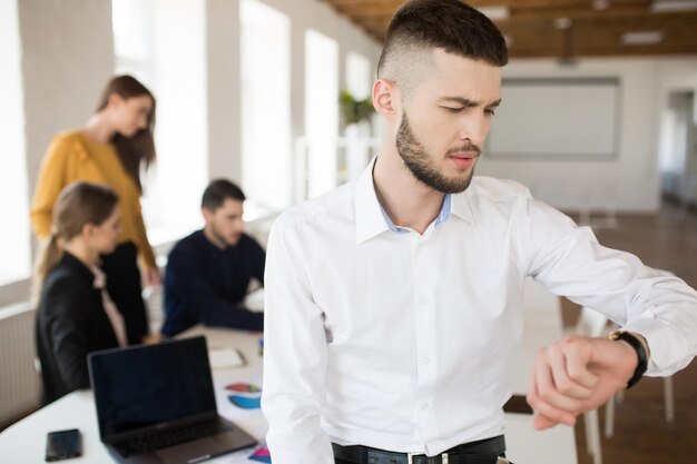 Jonge man met baard in wit overhemd die bedachtzaam op handhorloge kijkt terwijl hij tijd doorbrengt op kantoor met collega's op de achtergrond