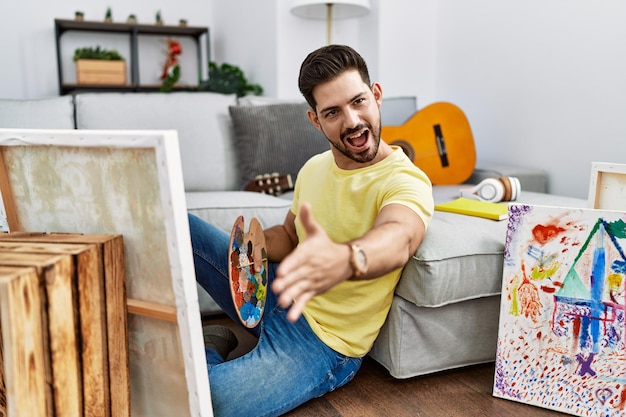 Jonge man met baard schilderdoek die thuis vriendelijk glimlacht en handdruk aanbiedt als begroeting en succesvol bedrijf verwelkomt