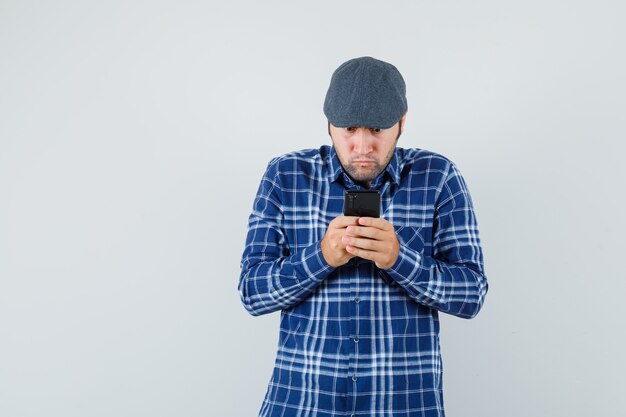 Jonge man met behulp van mobiele telefoon in shirt, pet en op zoek gericht, vooraanzicht.