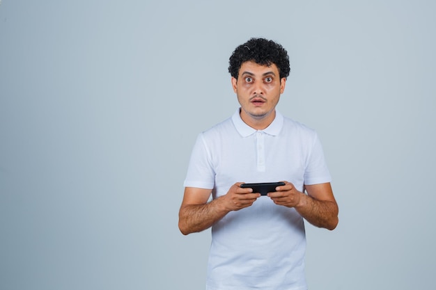 Jonge man met behulp van mobiele telefoon in wit t-shirt en op zoek verbaasd. vooraanzicht.