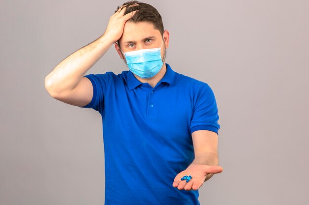 Jonge man met blauw poloshirt in medisch beschermend masker op zoek onwel en ziek staan met de hand op het hoofd lijden aan hoofdpijn met schillen in open hand over geïsoleerde oranje muur