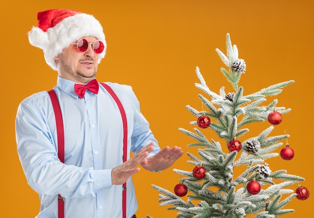 Jonge man met bretels vlinderdas in kerstmuts en rode bril die naast de kerstboom staat en ernaar kijkt met walgelijke uitdrukking hand in hand over oranje muur