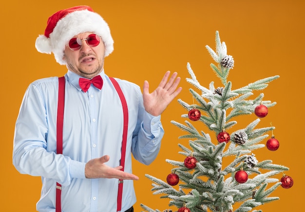 Gratis foto jonge man met bretels vlinderdas in kerstmuts en rode bril die naast de kerstboom staat en het presenteert met armen die geïrriteerd en ontevreden over oranje muur kijken