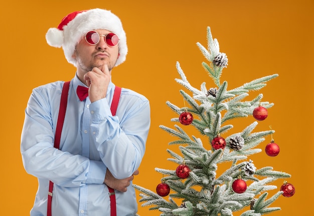 Jonge man met bretels vlinderdas in kerstmuts en rode bril kijken verbaasd staande in de buurt van de kerstboom over oranje muur