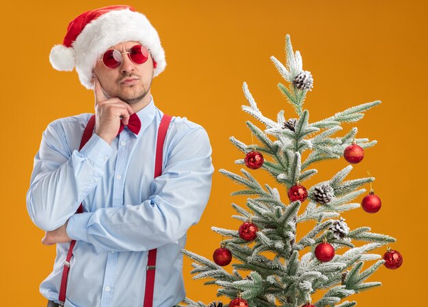 Gratis foto jonge man met bretels vlinderdas in kerstmuts en rode bril staan naast kerstboom met peinzende uitdrukking op gezicht denken over oranje muur